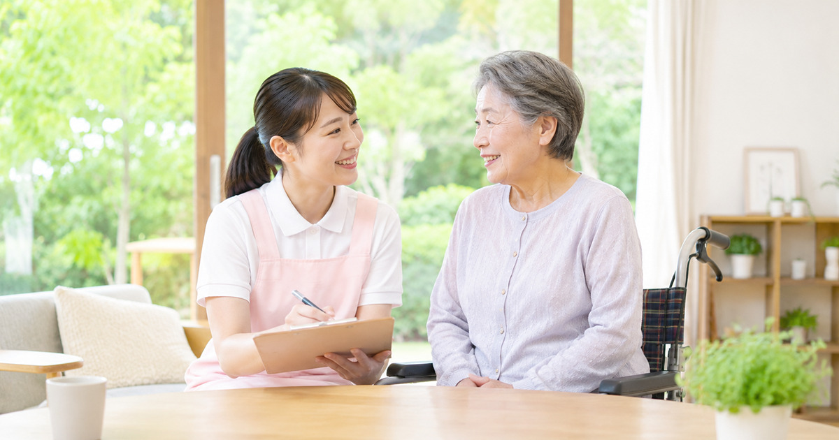 Caregiver in a pink apron sits with an elderly woman in a wheelchair, smiling and talking at a wooden table in a bright living room.