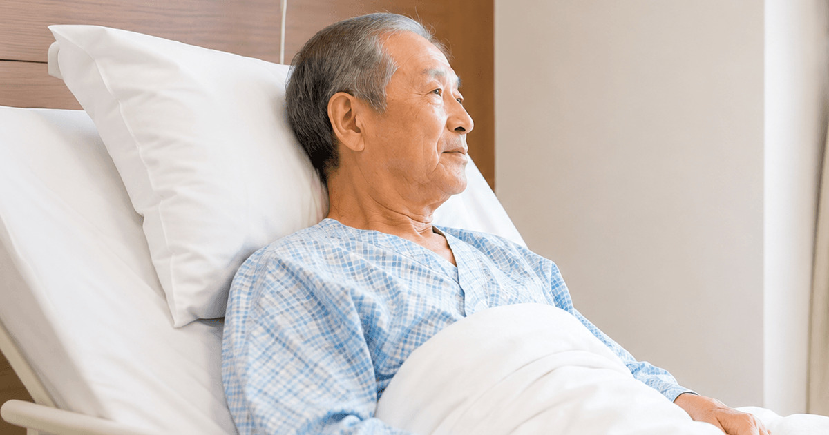 Older man in a blue hospital gown resting in bed with white pillows in a hospital room.
