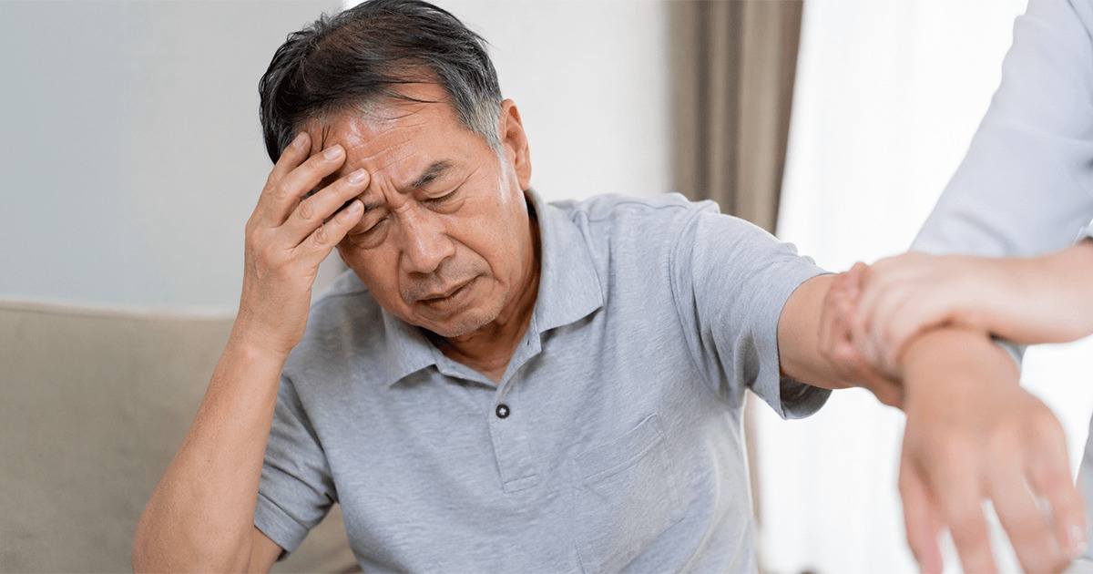 Older man sits on a couch, head in hand, showing pain or dizziness as a caregiver holds his arm to support him.