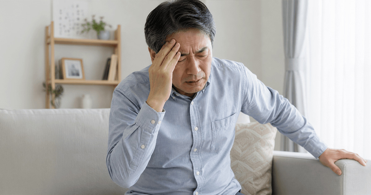 Man sitting on a living room sofa, hand on his forehead as if in headache or worry, with a neutral home setting behind him.