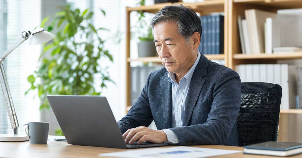 Senior man in a blazer typing on a laptop at a tidy office desk with a mug nearby and a bookshelf in the background